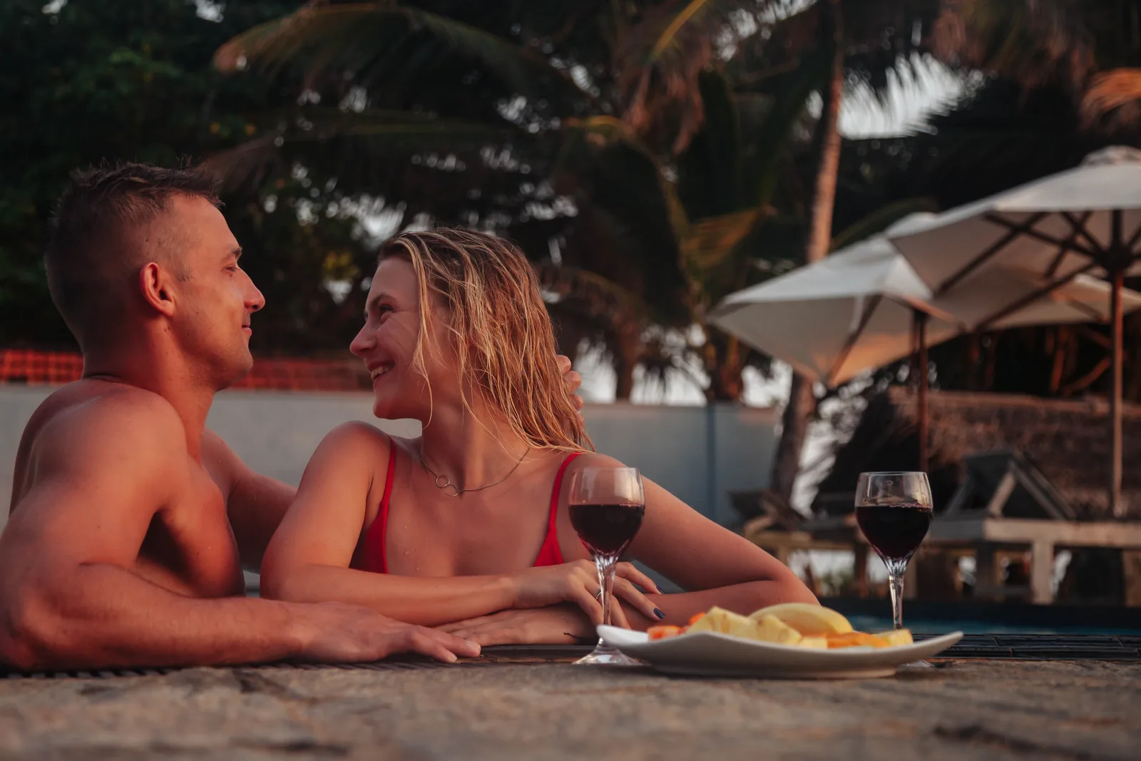 Adult couple sharing wine beside a tropical pool at sunset