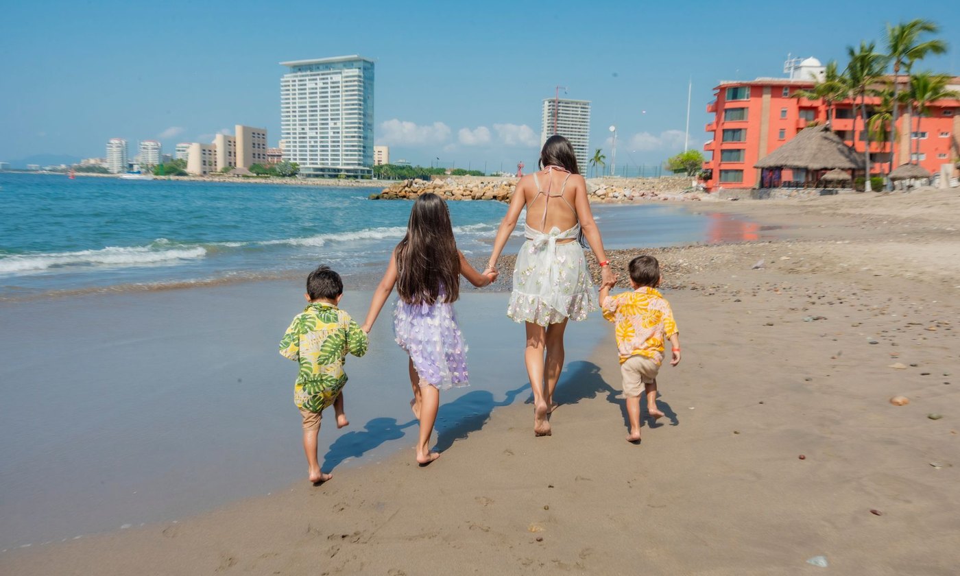 Mother and children walking along a sunny beach with a cityscape backdrop, enjoying family time.